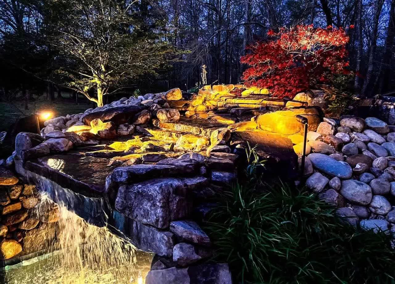 Landscape lighting illuminating a stone waterfall water feature at dusk